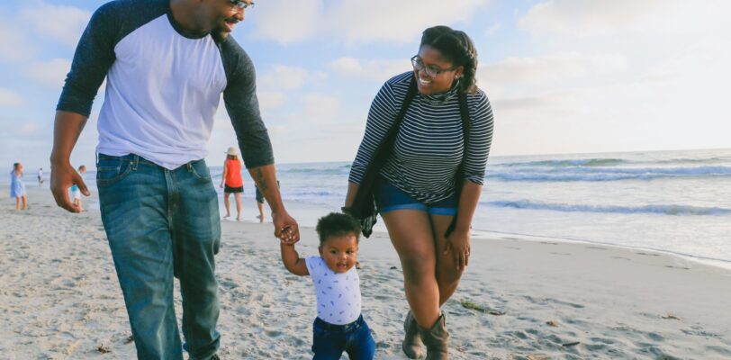 Man and woman holding the hands of a toddler while they are at the beach.